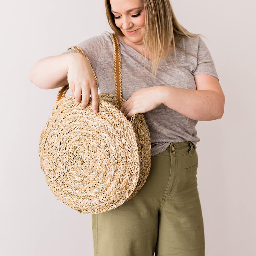 A person with blonde hair, wearing a gray t-shirt and olive green pants, is holding and looking into a round, woven straw bag with leather straps from The Bargain Sisters, against a plain, light-colored background.