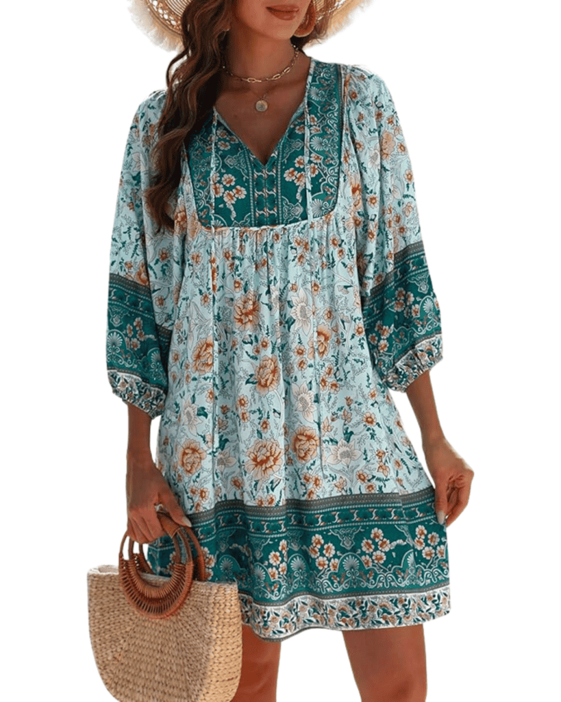 A woman in an affordable spring dress, featuring a floral pattern in green and orange hues. The dress boasts three-quarter sleeves and a V-neck. She complements her look with a straw handbag and chic sun hat, capturing the essence of effortless spring style.