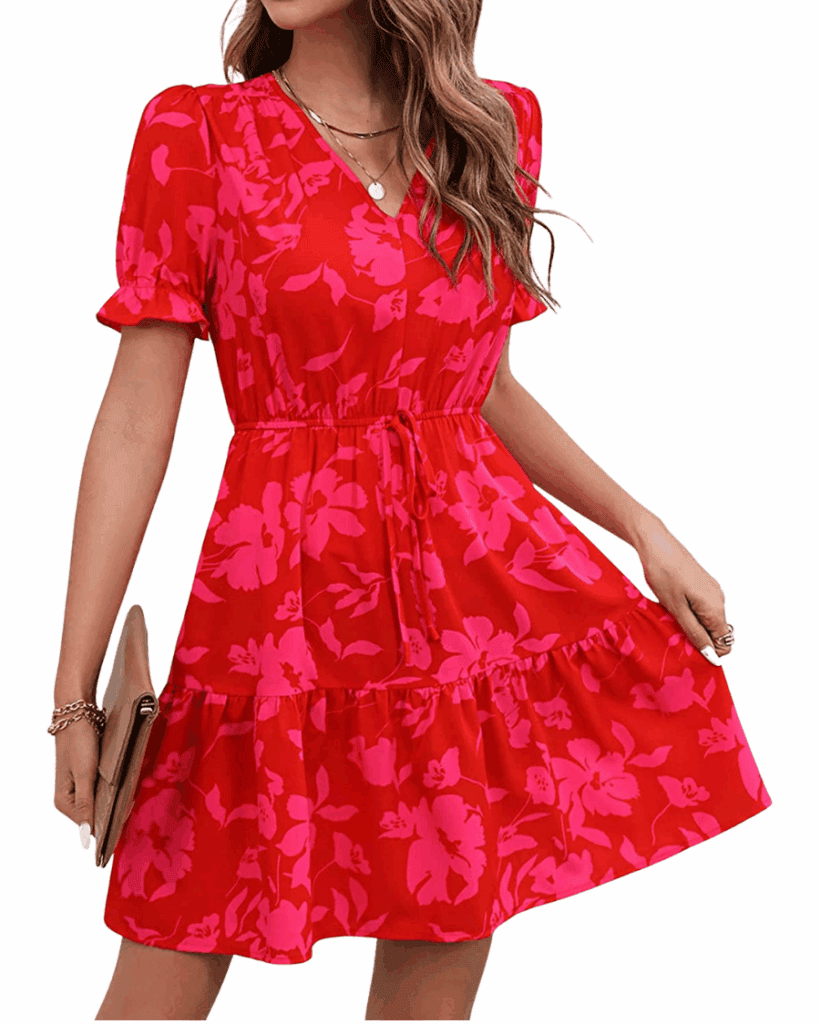 A woman wearing a short-sleeve, red dress with a pink floral pattern, a V-neckline, a cinched waist with a tie, and a ruffled hem. She is holding a tan clutch and wearing bracelets.