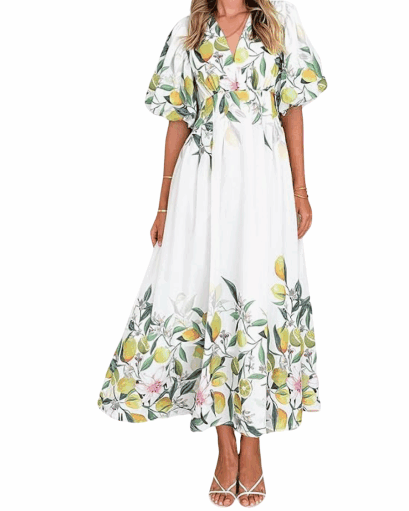 A woman wearing a white, flowy maxi dress with green and yellow lemon print, puffed short sleeves, a V-neckline, and white heeled sandals.