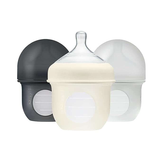 Three baby bottles with wide necks and rounded caps are shown in black, white, and off-white. The front bottle features a clear silicone nipple and measurement markings—perfect for stocking up during Monday Deals!.