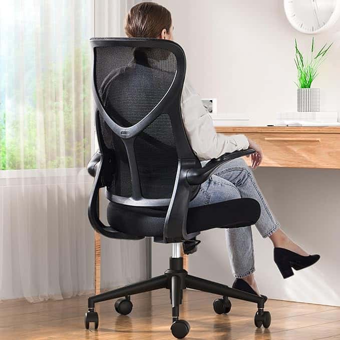 A person sits at a wooden desk in front of a window, enjoying their high-back ergonomic office chair with a black mesh design—perfect for browsing Tuesday Deals. The person has light brown hair and is wearing a light jacket, jeans, and dark shoes.