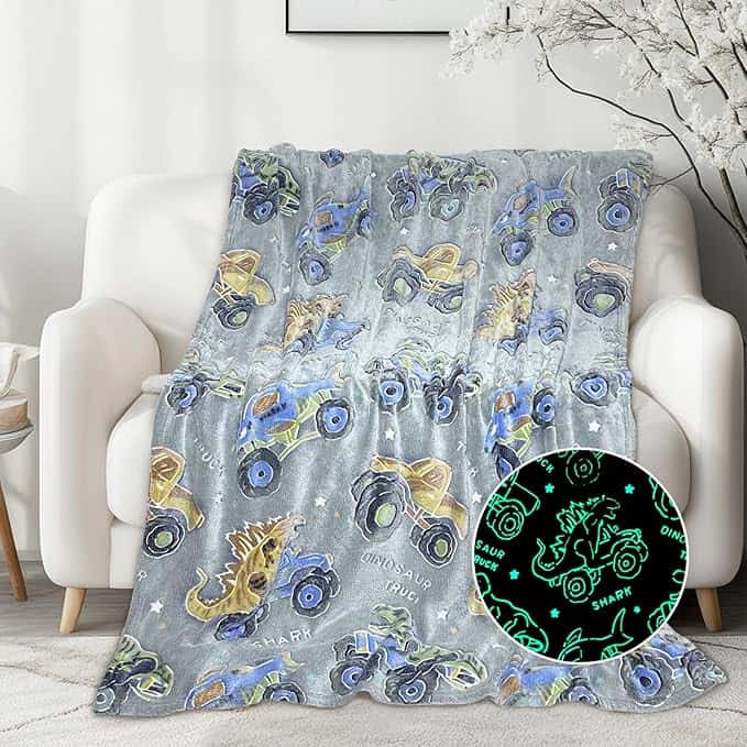 A gray blanket with colorful monster trucks and dinosaurs draped over a white sofa. An inset shows the blanket glowing in the dark with green outlines and words like "DINOSAUR" and "SHARK." Perfect for cozy Tuesday Deals!.