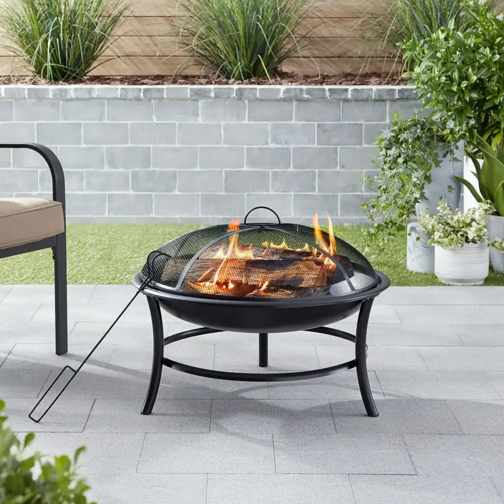 A black metal fire pit with burning logs and a mesh cover stands on a gray patio. Perfect for cozy gatherings, this setup is ideal for enjoying Friday Deals. Green plants, a cushioned bench, and a stone wall complete the scene.