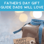 A wrapped gift with a blue ribbon and a tag that says "Happy Father's Day," with text above reading "Father’s Day Gift Guide Dads Will Love." Blurred lights are in the background.