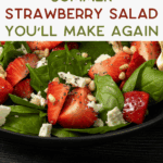 A fresh summer salad with spinach, sliced strawberries, crumbled cheese, pine nuts, and poppy seeds in a black bowl. Text overlay reads, “Summer Strawberry Salad You’ll Make Again.”.