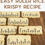 Rectangular Rice Krispy treats decorated with black icing lines to look like rulers are arranged on a wooden board. The text above reads “Easy Ruler Rice Krispy Recipe.” Two glasses of milk are in the background.