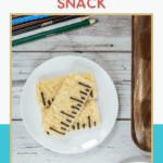 A white plate holds two rectangular Rice Krispy treats decorated with black icing lines to resemble rulers. Colored pencils and a wooden tray are nearby on a white wooden table.
