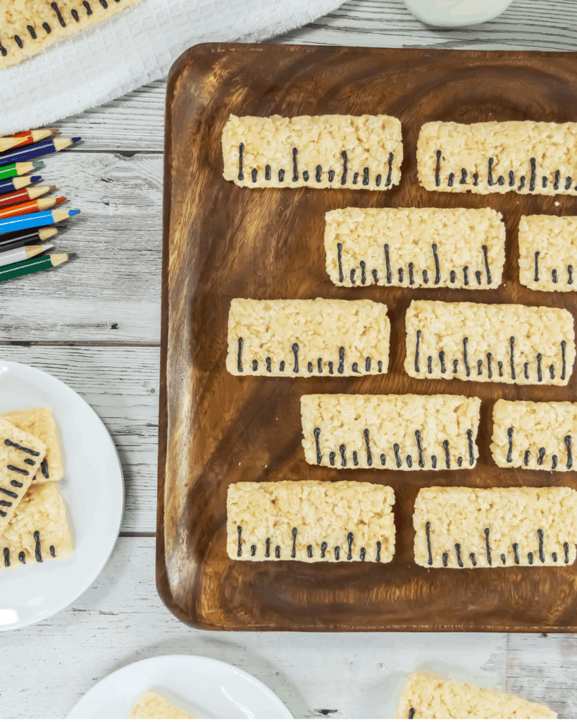 Rectangular Rice Krispies treats, made using a precise ruler rice krispy recipe, are arranged on a wooden tray and decorated with black icing to resemble piano keys. Plates of more treats and colored pencils sit nearby on a white wooden table.