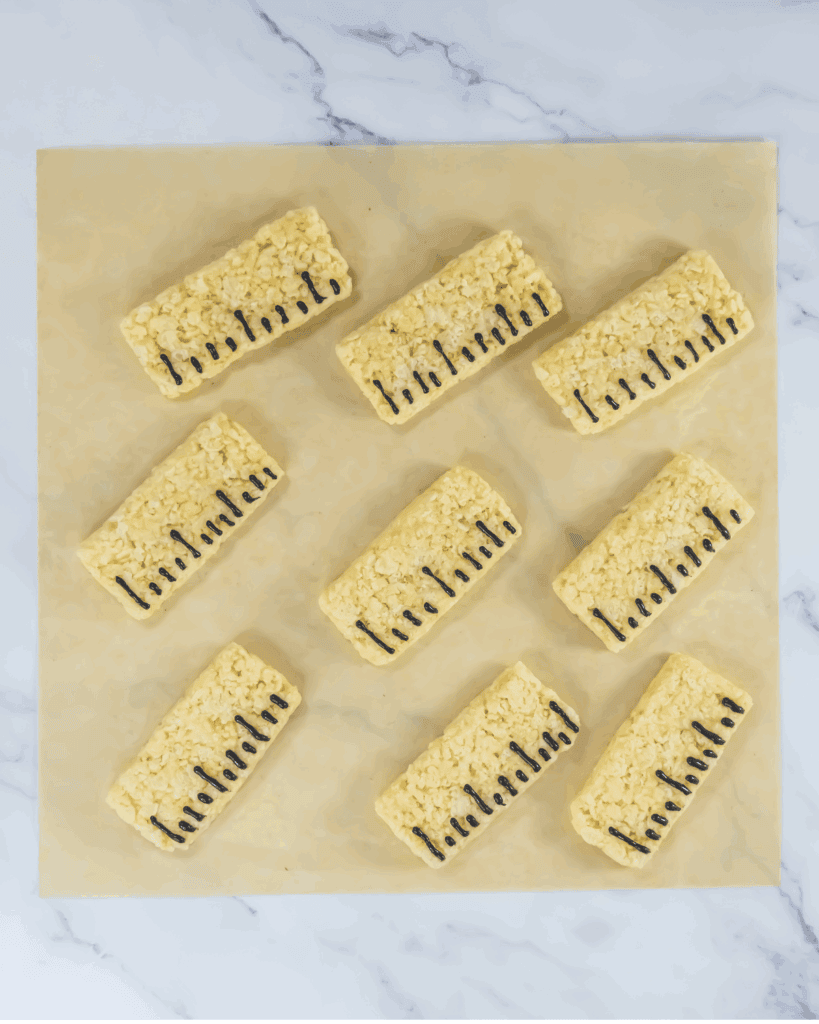 Eight rectangular Rice Krispies treats, made with a classic ruler rice krispy recipe, are arranged on parchment paper. Each treat features a line of black icing resembling piano keys. The background is a white marble surface.