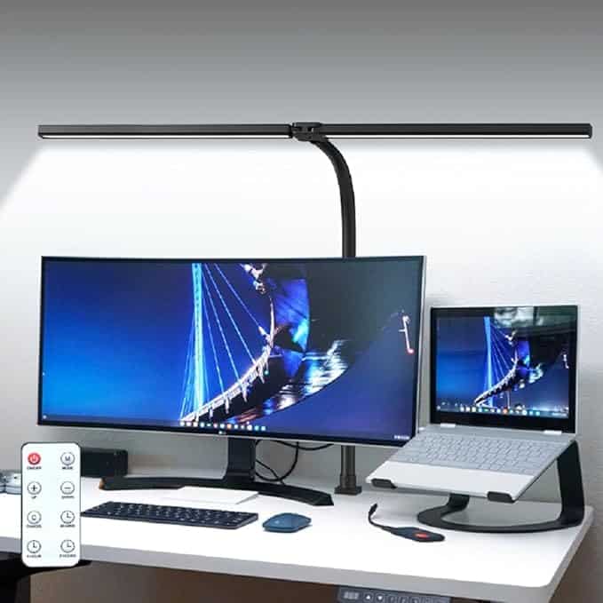 A modern desk setup with a curved monitor and open laptop, both displaying a blue-lit bridge. A long, slim desk lamp illuminates the area—perfect for catching Monday Deals—with a remote control, keyboard, and mouse on the clean white desk.