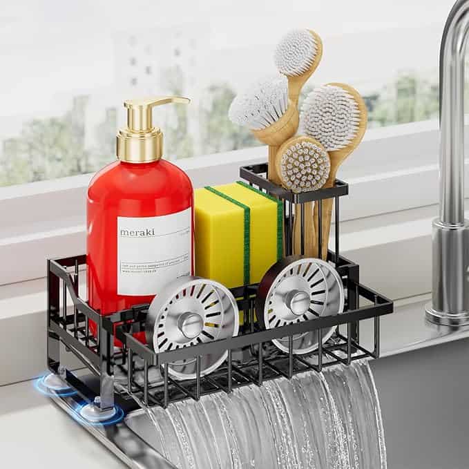 A black sink caddy holding a red soap dispenser, two yellow sponges, two wooden brushes, and two silver sink strainers sits on a sink edge as water flows beneath it, with Tuesday Deals shining through the window in the background.