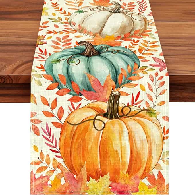 A table runner with watercolor-style pumpkins in orange, teal, and white, surrounded by colorful autumn leaves and branches on a white background, displayed on a wooden table—perfect for refreshing your décor with Monday Deals.