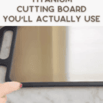 A hand points to a sleek titanium cutting board with a black handle on a marble countertop. Text above reads: "Titanium cutting board you’ll actually use.