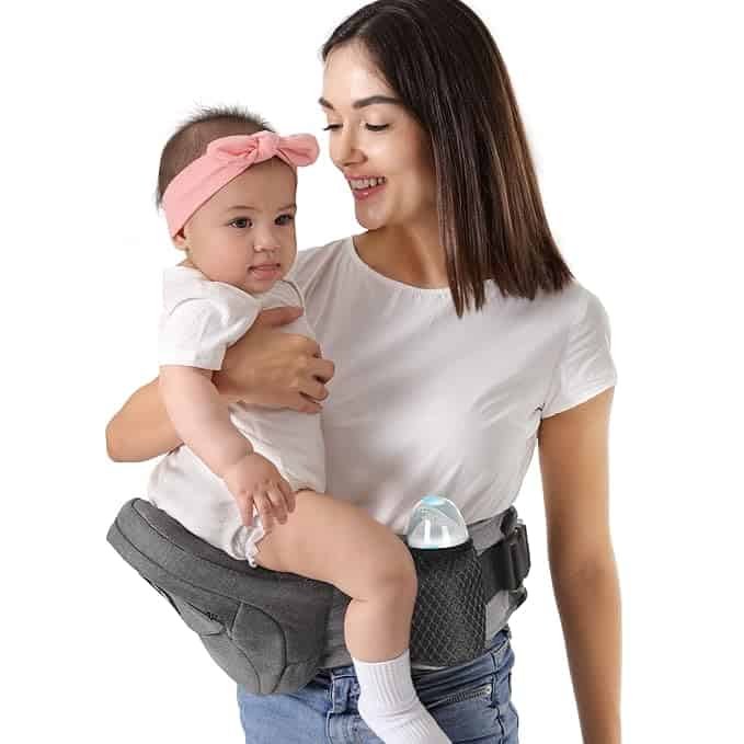 A smiling woman in a white shirt holds a baby with a pink headband on a gray hip seat carrier, featuring a baby bottle in the side pocket. Enjoy this moment and grab exclusive Monday Deals! The background is white.