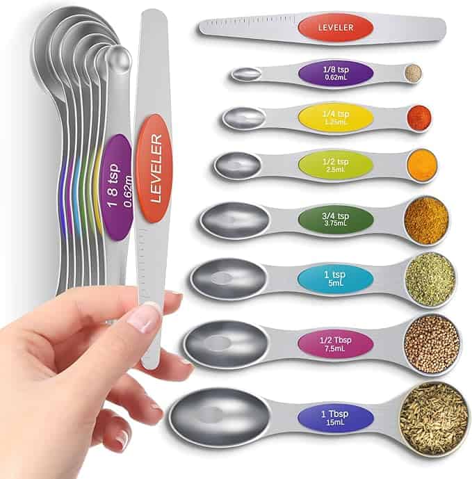 A set of stainless steel measuring spoons with colorful labels, each marked from 1/8 tsp to 1 Tbsp, plus a handy leveler tool. Some spoons filled with spices. A hand holds the leveler—perfect for your kitchen and Thursday Deals!.