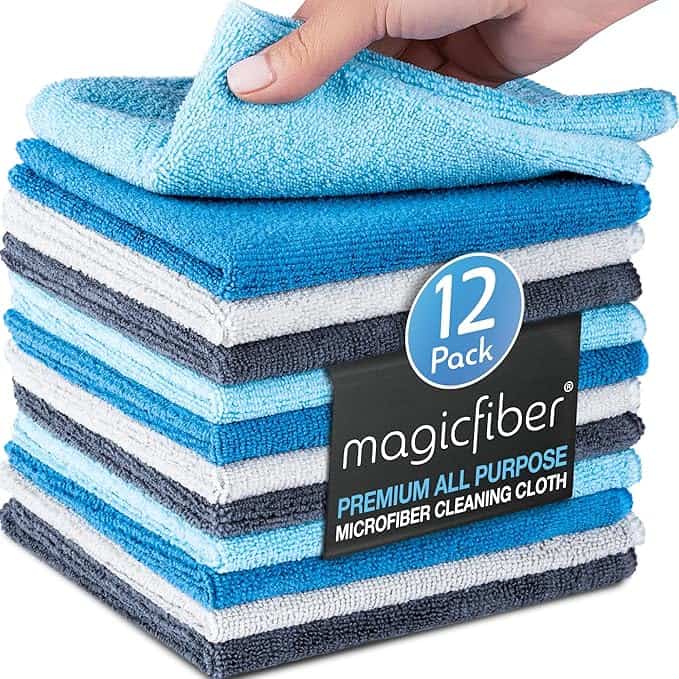 A hand lifts a light blue microfiber cleaning cloth from a stack of folded cloths in shades of blue, gray, and white. The packaging reads "12 Pack, magicfiber, Premium All Purpose Microfiber Cleaning Cloth." Perfect for Monday Deals!.