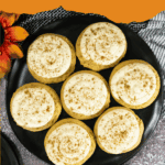 A black plate holds seven pumpkin cookies topped with swirled caramel frosting and a sprinkle of spice. A bowl of caramel sauce and a flower are beside the plate. Text at the top reads "Easy Caramel Pumpkin Cookies.