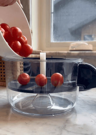 A hand pours cherry tomatoes from a white bowl into a transparent kitchen container with a metal blade inside—an automatic peeler sits nearby on the marble countertop near a window.