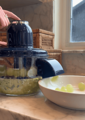 A person uses a juicer to crush green grapes on a kitchen counter, with some whole grapes falling into a white dish beside the juicer. An automatic peeler sits nearby, while wicker boxes and a window are in the background.