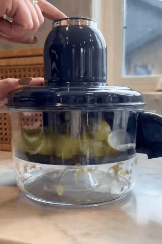 A person presses a button on top of an electric food chopper with chopped vegetables swirling inside a glass bowl, while an automatic peeler rests nearby on the marble countertop near a window.