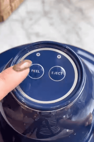 A hand with painted nails is pressing the "PEEL" button on a blue automatic peeler. Next to it is an "EJECT" button, both set on a circular control panel.