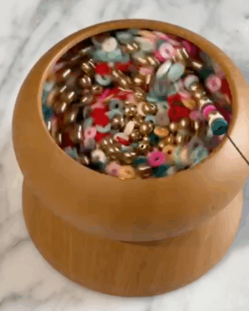 A wooden bead spinner filled with colorful beads spins rapidly, causing the beads to swirl around inside the bowl. A needle with a thread is picking up beads from the spinner.