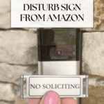 A hand with pink nail polish holds a small sign reading "NO SOLICITING" in front of a video doorbell mounted on a stone wall. Text above reads: "Cute Do Not Disturb Sign from Amazon—perfect for making mealtime easier and uninterrupted.