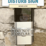 A video doorbell mounted on a stone wall has a "Do Not Disturb" sign attached to it. Text above reads, "Review: Modern Do Not Disturb Sign Moms Love—Easier Mealtime," with a website link at the bottom.
