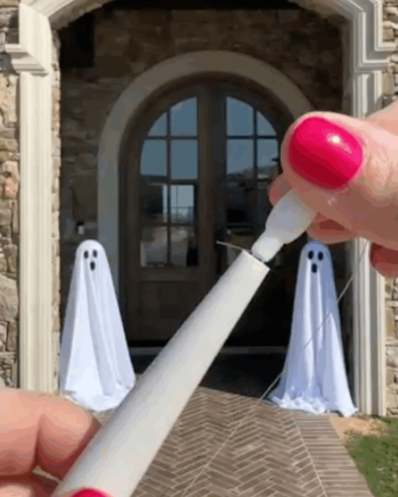 Close-up of hands with pink nail polish assembling a white decoration stick, while two ghost decorations made from white sheets stand outside near a stone entrance and an air conditioner cover in the background.
