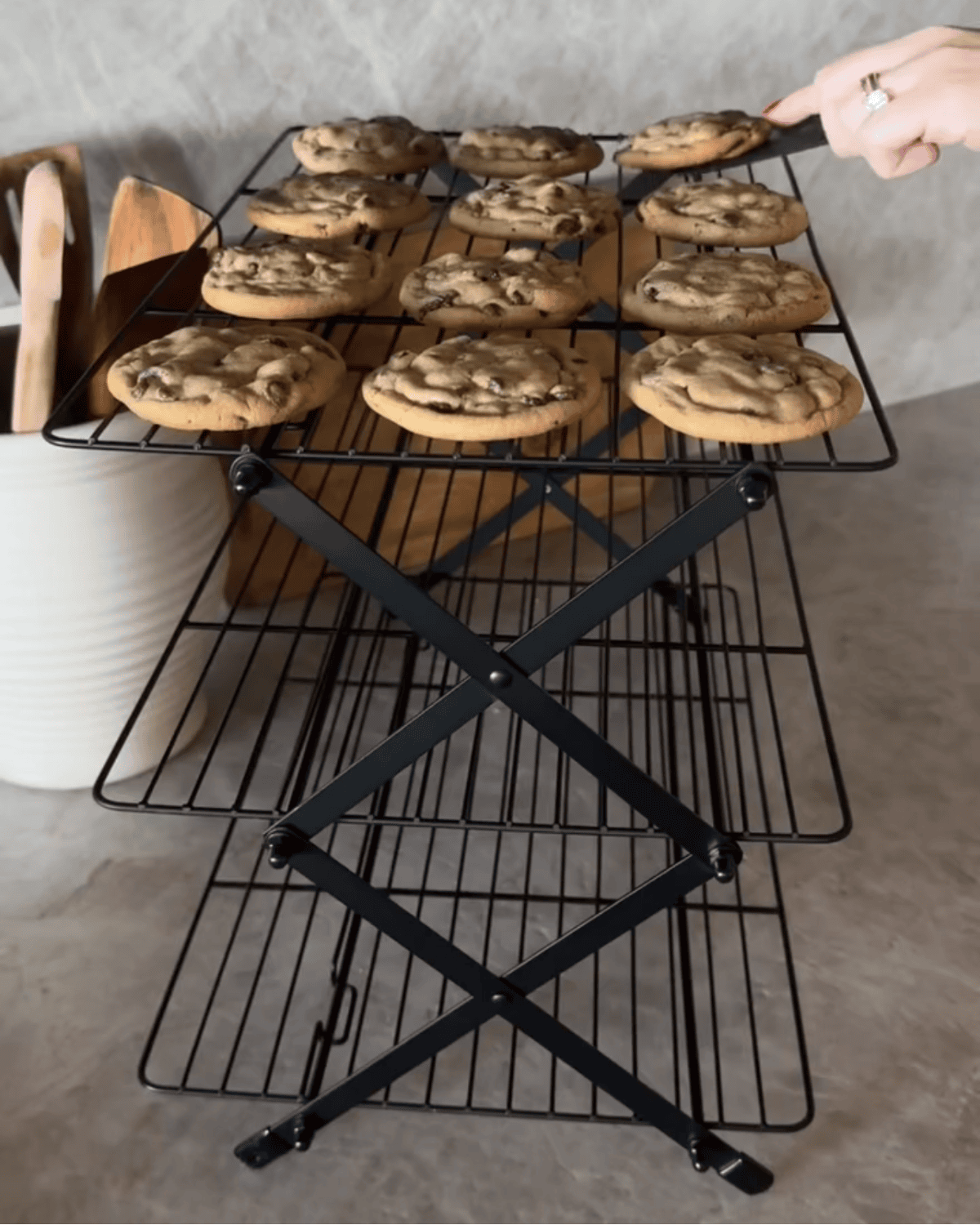 A 3 tier cooling rack holds freshly baked chocolate chip cookies, with a hand using a spatula to lift one. A white container filled with wooden utensils sits beside the rack on the kitchen countertop.