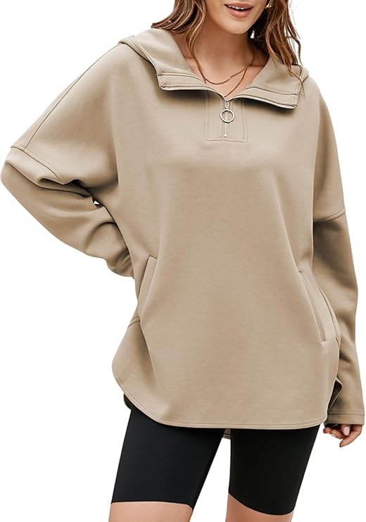 A woman wearing a loose, beige pullover sweatshirt with a wide collar and front pocket, paired with black biker shorts. She has long, wavy hair and is smiling—perfect for comfy style or scoring Thursday Deals.