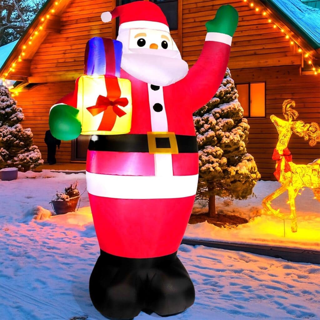A large inflatable Santa Claus holding a gift stands on snow in front of a wooden house decorated with lights. Snow-covered trees and a glowing reindeer decoration are visible in the background.