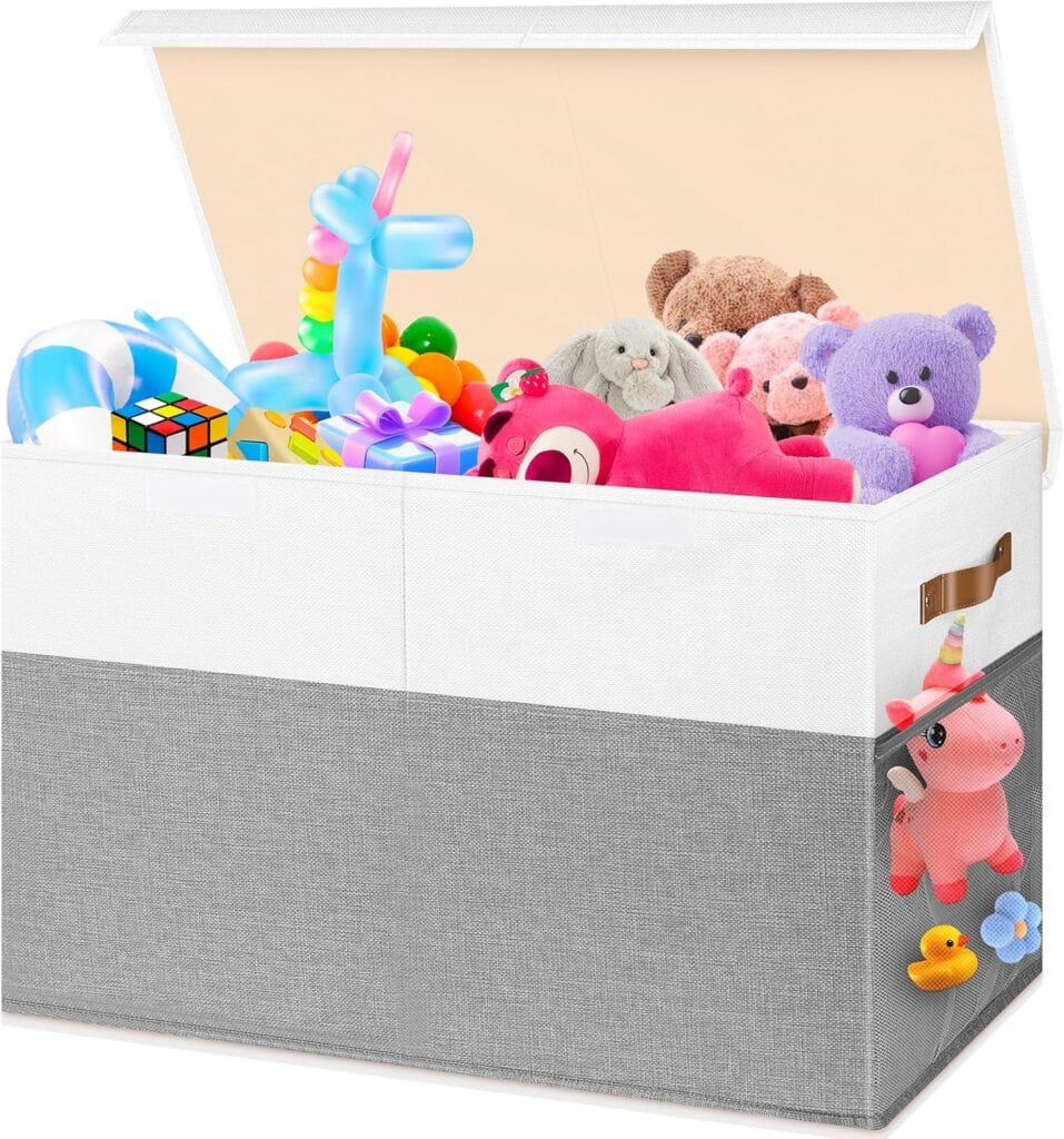 A large, white and gray fabric toy storage box with an open lid is filled with colorful toys, including stuffed animals, a Rubik’s Cube, and a blue balloon animal. A pink unicorn plush is attached to the side pocket.