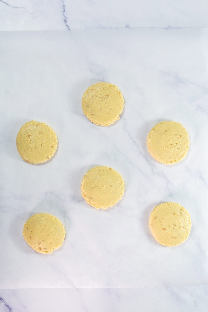 Six round, unbaked Twix cookies dough discs are evenly spaced on a sheet of parchment paper atop a light marble surface.