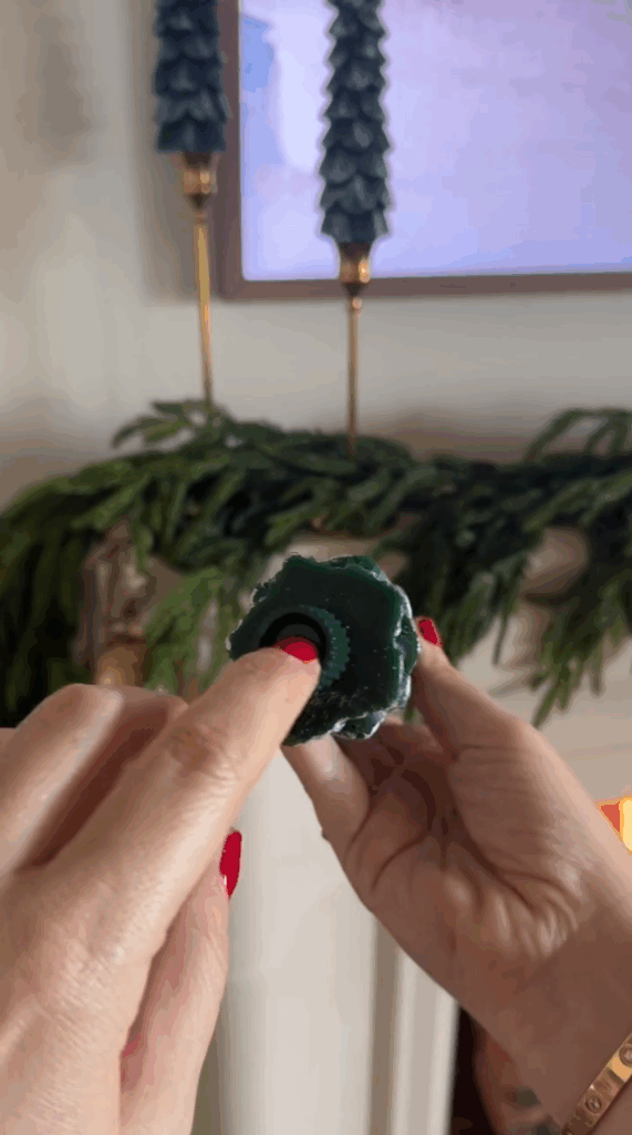 A person with red nail polish holds a green, flower-shaped candle with a black center. In the background, green garlands and two dark green tapered Christmas candles decorate the mantel.