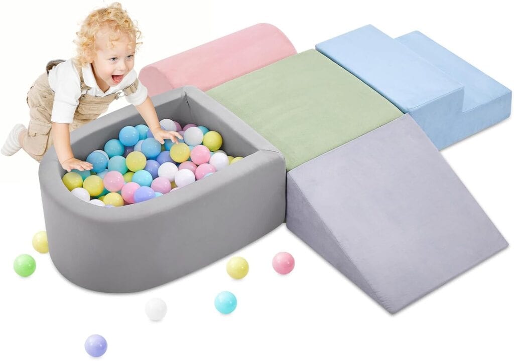 A smiling toddler climbs into a gray foam ball pit filled with colorful balls, next to soft, pastel-colored foam climbing blocks and steps. Some balls are scattered outside the pit.