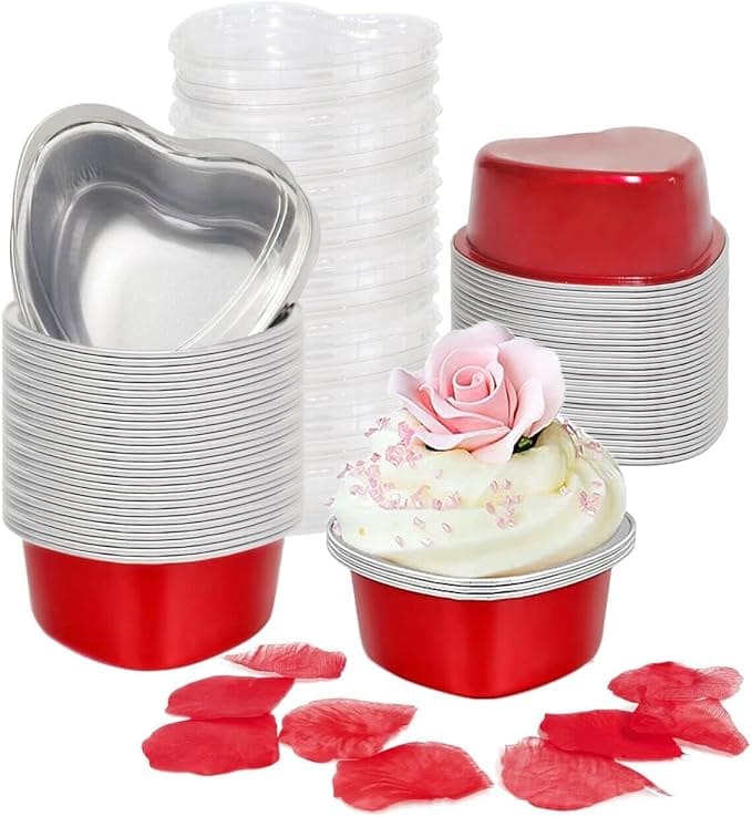 Stacks of red and silver heart-shaped aluminum baking cups with clear plastic lids, a cupcake topped with a pink rose, and red rose petals scattered in front.