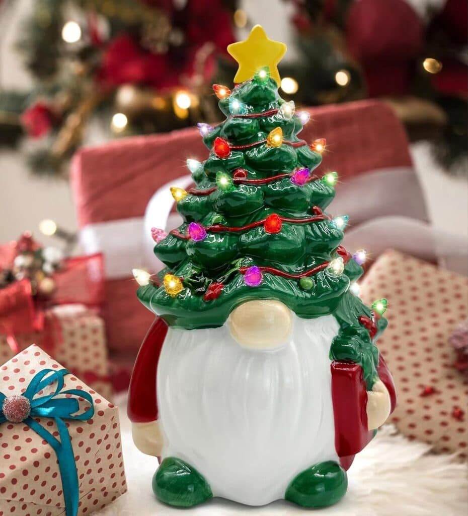 A festive gnome figurine with a Christmas tree hat decorated with colorful lights and a yellow star stands among wrapped presents, with a decorated tree in the blurred background.