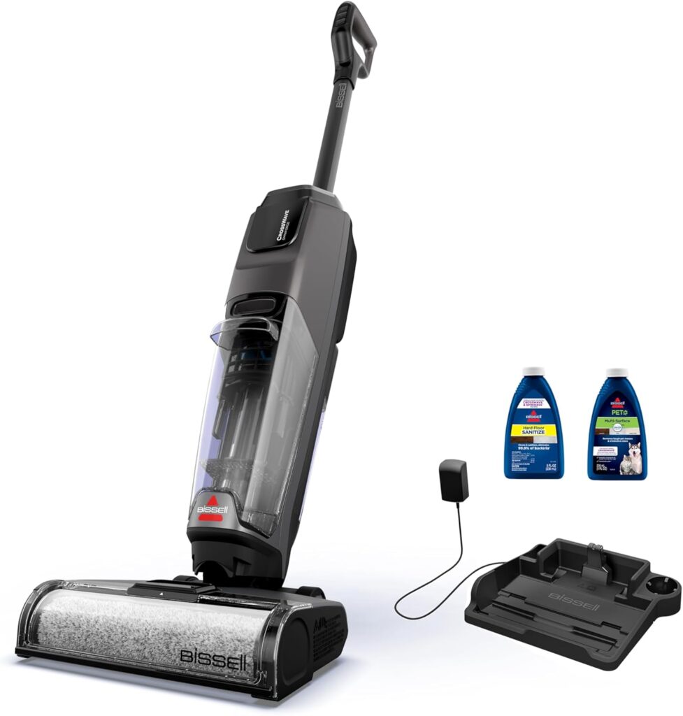 A black and gray BISSELL wet and dry vacuum cleaner stands upright next to a charging base, charger, and two bottles of cleaning solution, all displayed on a white background.