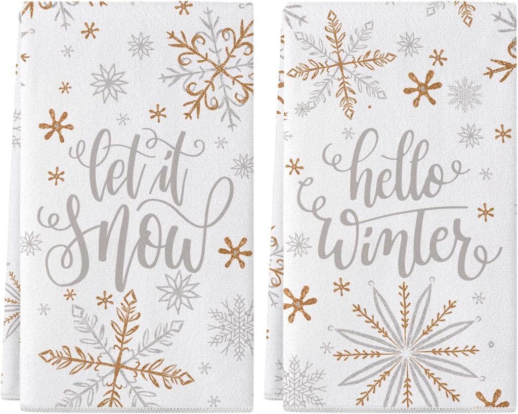 Two white towels decorated with silver and gold snowflakes. One towel has the words "let it snow" in script, and the other says "hello winter," both surrounded by various snowflake patterns.