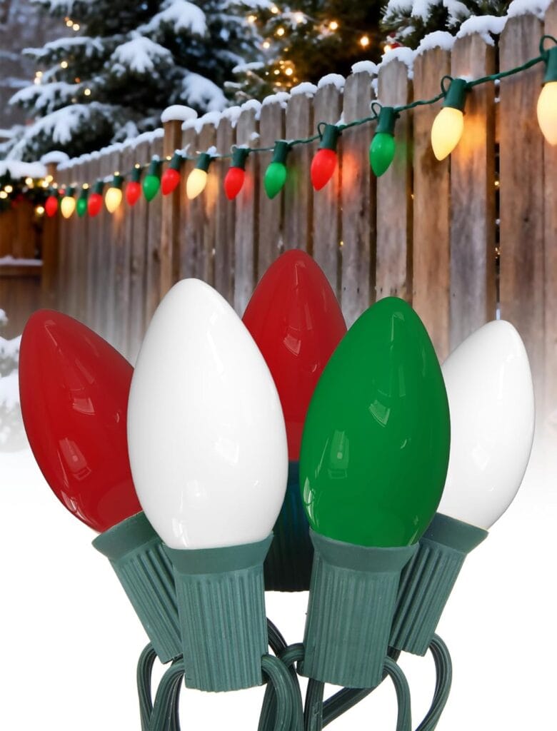 A close-up of colorful Christmas lights—red, white, and green bulbs—on a green cord, with a snowy wooden fence decorated with glowing lights and snow-covered trees in the background.