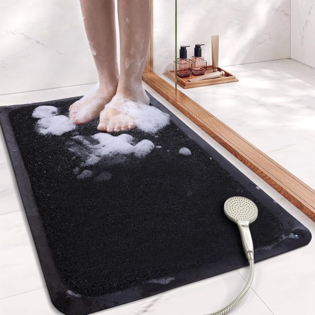 A person stands barefoot on a black bath mat covered with soap suds in a bathroom. A handheld shower head rests on the mat, and soap bottles are visible on a wooden tray near a glass shower wall.