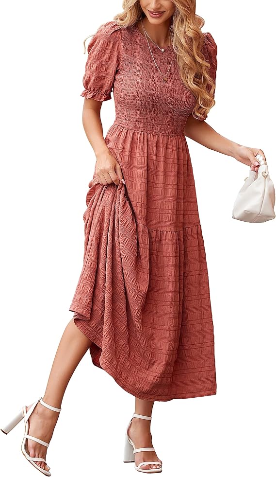 A woman in a textured, short-sleeve, rust-colored midi dress holds the skirt with one hand and a small white handbag in the other, wearing white high-heeled sandals and layered necklaces.