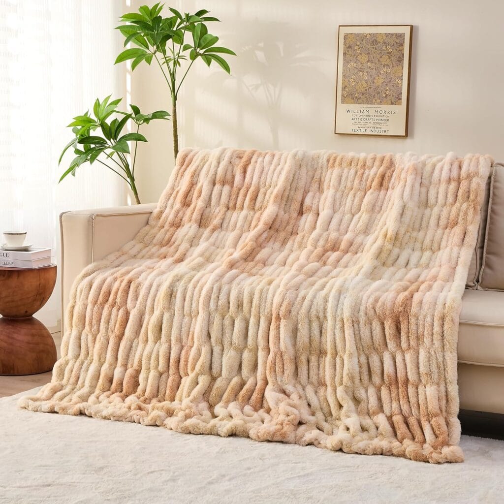 A plush, cream and light peach Faux Fur Throw Blanket draped over a beige sofa in a bright living room with a potted plant, framed wall art, and wooden side table. Sunlight streams through sheer curtains. Enjoy 50% off for a limited time!.