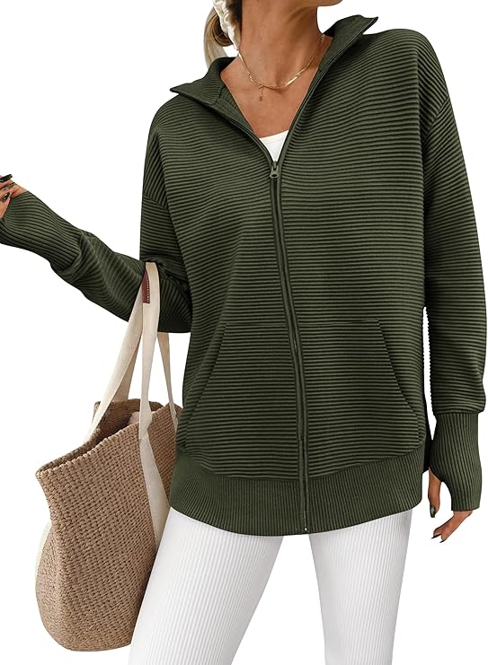 A person wearing a ribbed, olive green zip-up jacket over a white top and white pants, holding a large beige woven tote bag. The image is cropped at the neck, and the background is white.