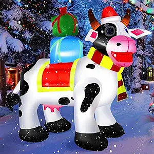 An inflatable cow with black spots, wearing a red Santa hat and yellow scarf, stands in the snow with holiday presents on its back. Snow-covered trees and festive lights are in the background.