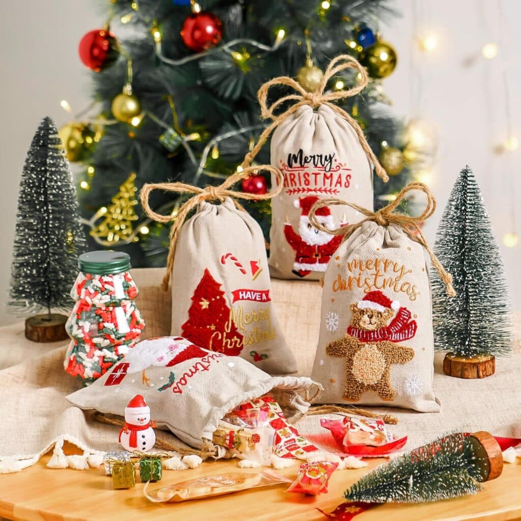 Christmas gift bags with festive designs sit on a table alongside a jar of candy, small Christmas trees, and holiday treats. A decorated Christmas tree with ornaments and lights is in the background.