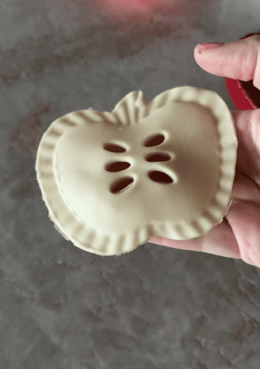 A hand holding an unbaked pastry shaped like an apple, with small cutouts resembling apple seeds and crimped edges, crafted using pie molds.