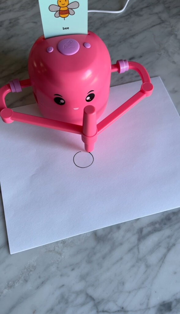 A pink drawing robot with a smiling face draws a black circle on white paper placed on a gray marble surface. A card labeled "bee" is slotted on top of the kids robot.
